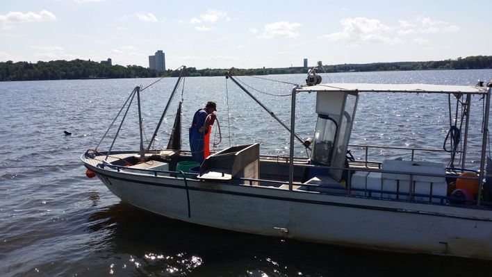 Bilden visar en fiskebåt i Mälaren. På båten finns en man, fiskaren, som håller på och lägger ut fisknät.